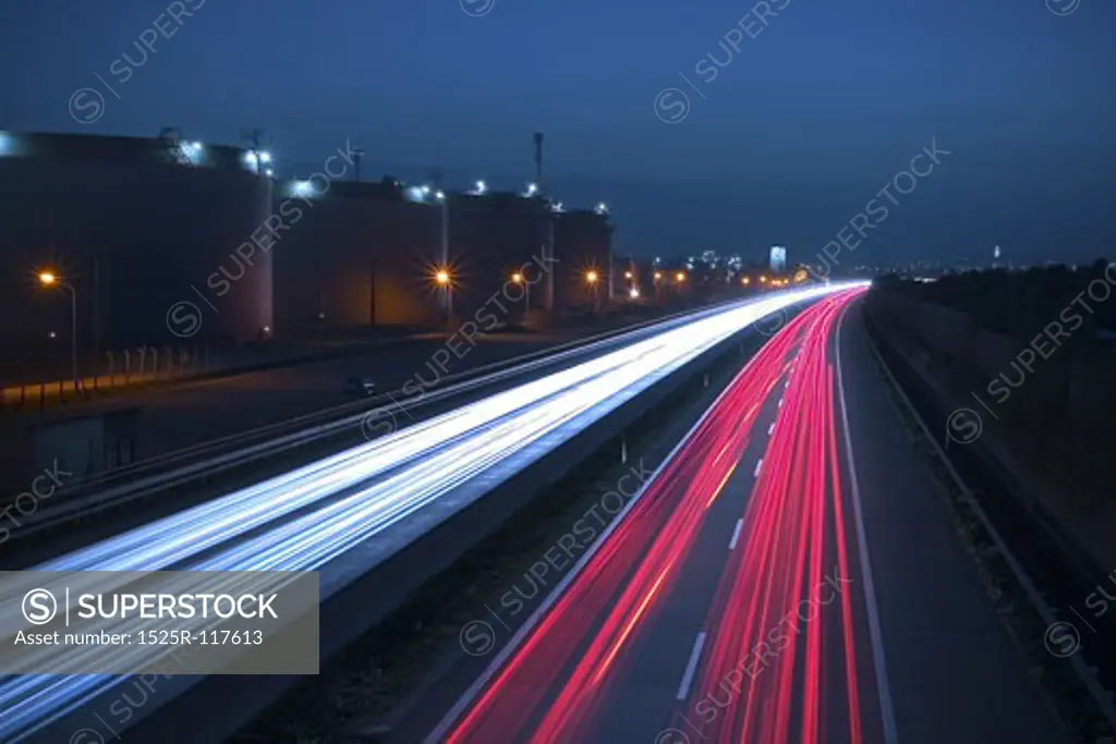 Heavy traffic on a highway. Due to the long exposure time the front and rear lamps of the cars are forimg a white and a red snake of light. There are huge storage tanks on the left side.