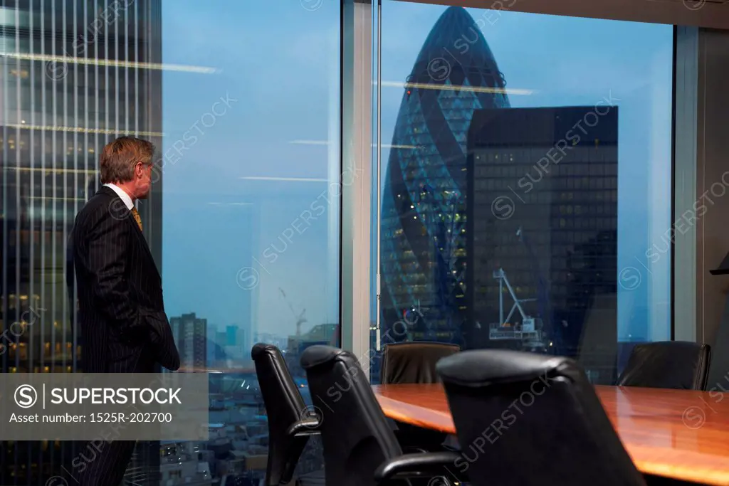 Senior business man standing in boardroom alone looking out of window of view of city