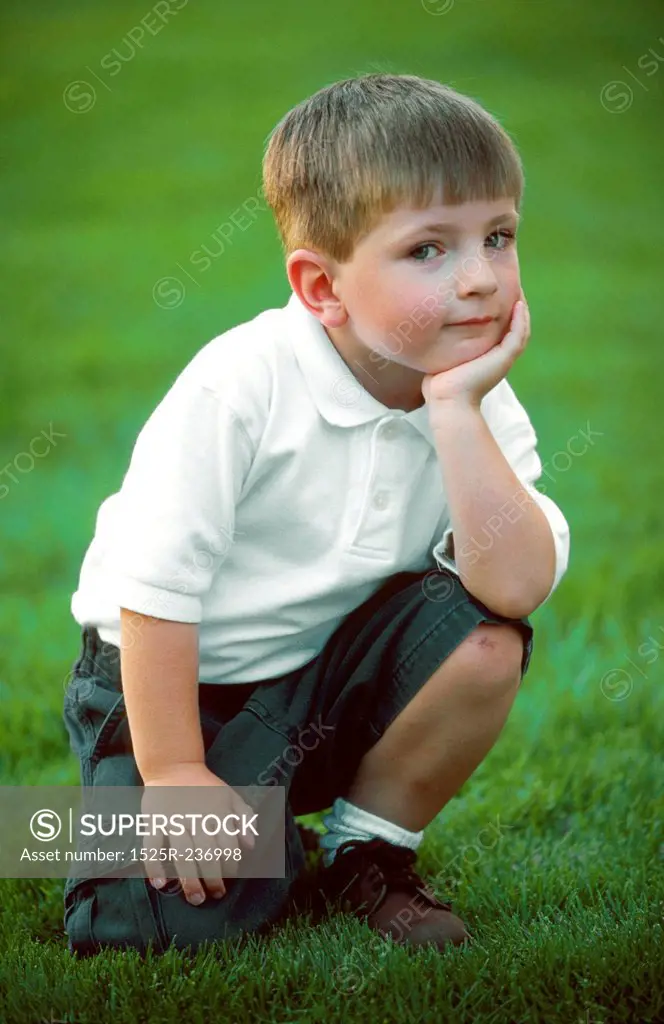 Young Caucasian Boy Kneeling In Green Grass And Smiling At You