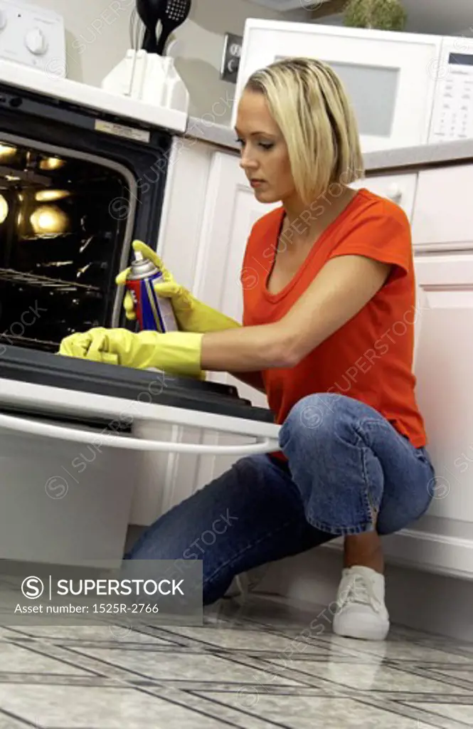 A young woman wearing kitchen mittens, cleaning an oven with oven spray