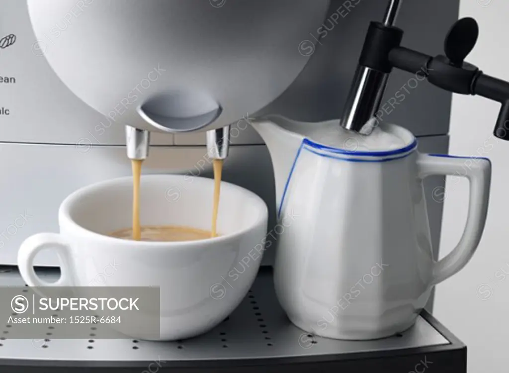 Cappuccino pouring from a coffee maker into a cup next to a kettle of steamed milk