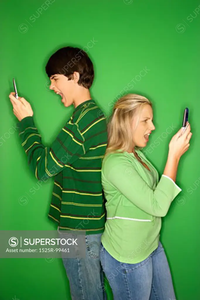 Portrait of Caucasian teen boy and girl on cellphones standing with backs to eachother against green background.
