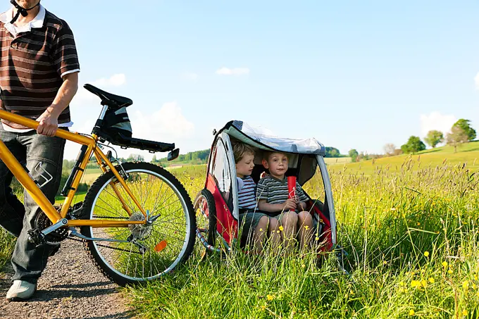 Dad driving his two children on a weekend excursion with bikes on a summer day in beautiful landscape, for safety and protection they are sitting in a bike trailer