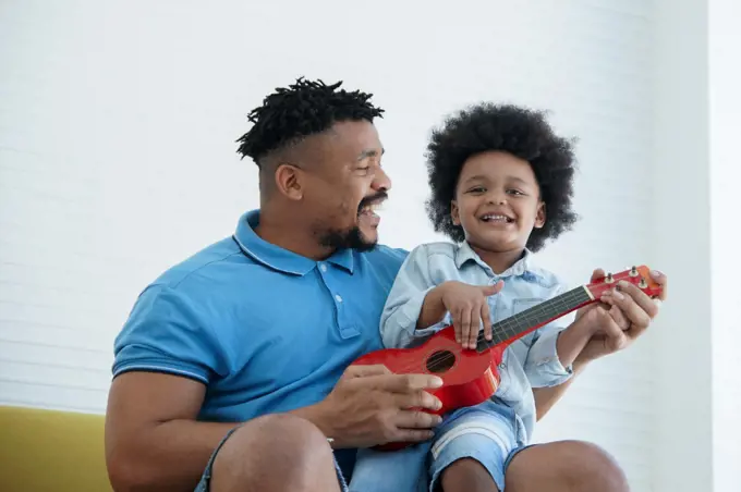 African American father and little son singing and playing ukulele together. Dad teaching his boy play a small red ukulele on sofa at home