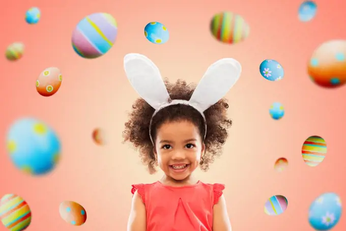 easter, holidays and childhood concept - happy little african american girl wearing bunny ears headband over living coral background and colored eggs. happy little girl wearing easter bunny ears