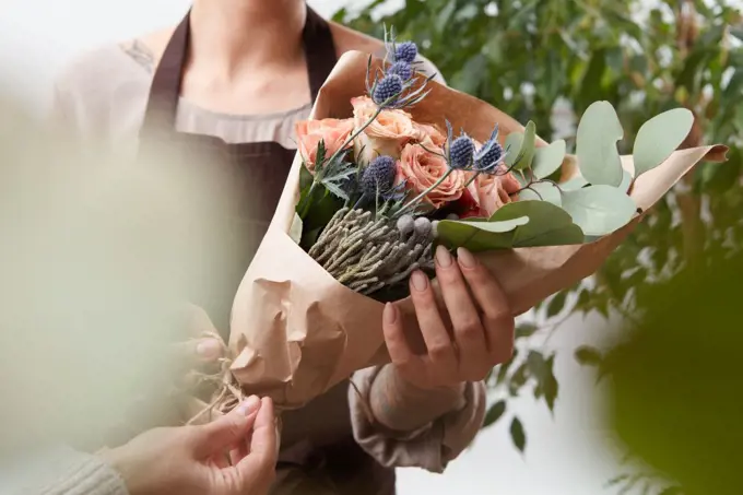 Bouquet of fresh fragrant roses living coral color in a paper with blured green leaf background. Concept of Mother Day.. Close-up of roses flowers bouquet in a woman&rsquo;s hands on a blured green leaf background.