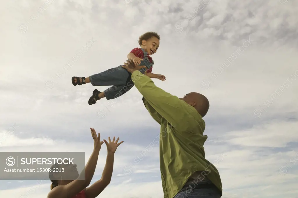 African-American father lifts his son into a cloudy white sky, with mother close by.  