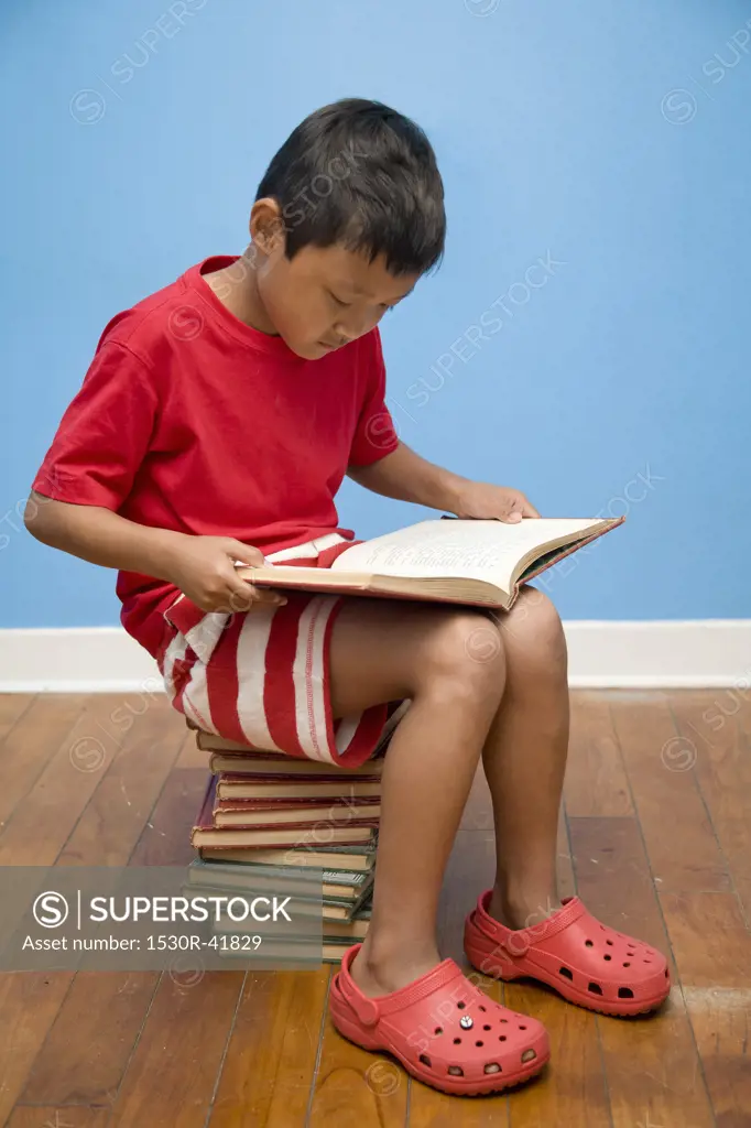Boy reading while seated on stack of books,