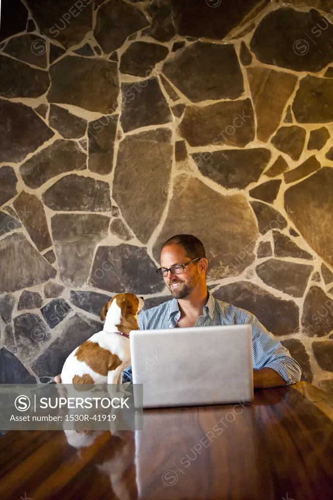 Man seated at long table with laptop and dog   