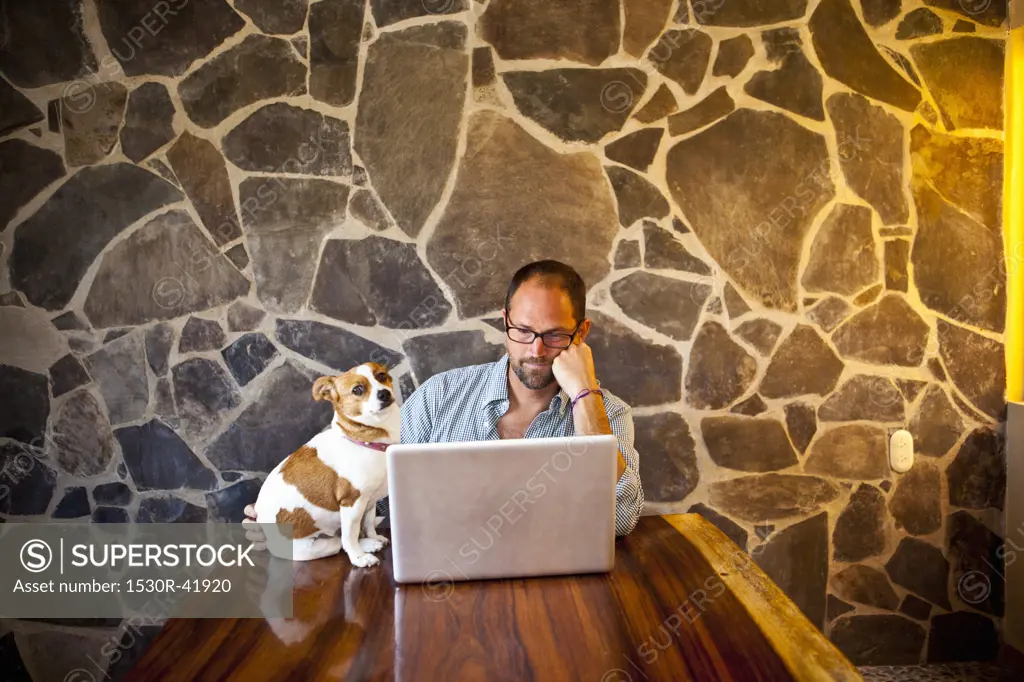 Man seated at long table with laptop and dog