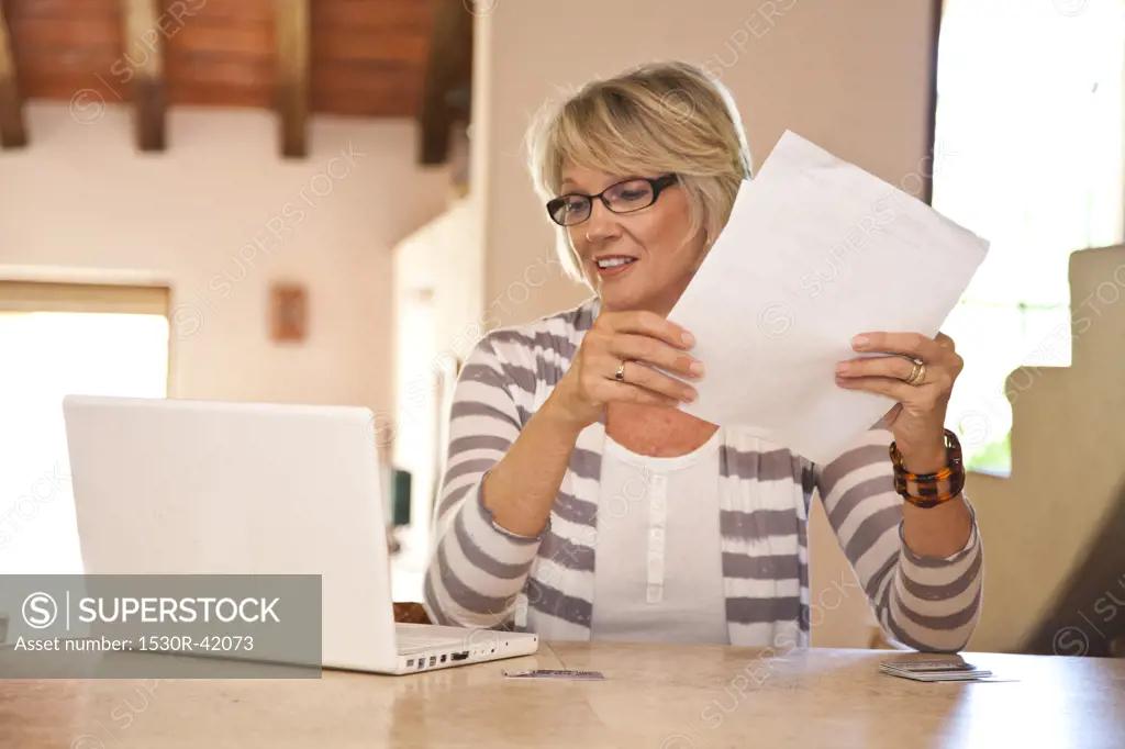 Woman at home office holding bills and looking at laptop,