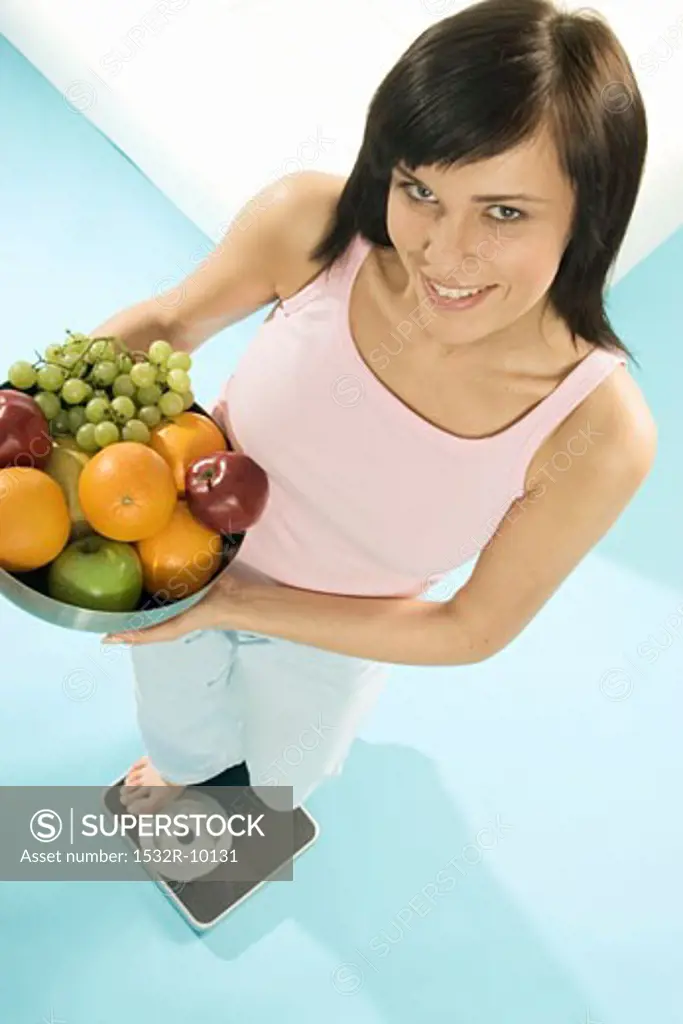 Young woman standing on scales with bowl of fruit
