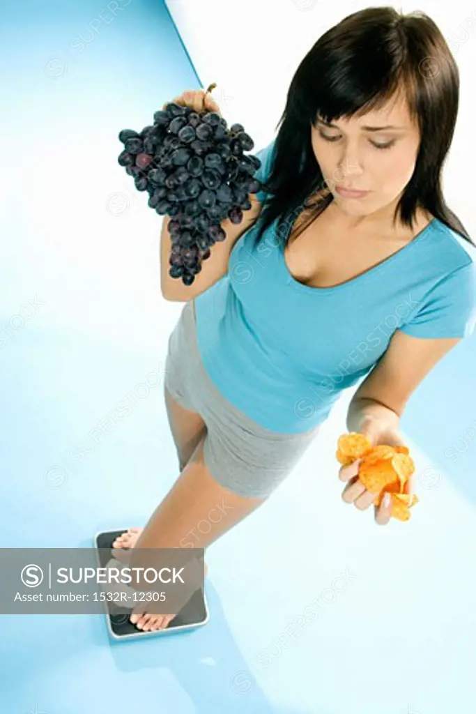 Young woman standing on scales with crisps and grapes