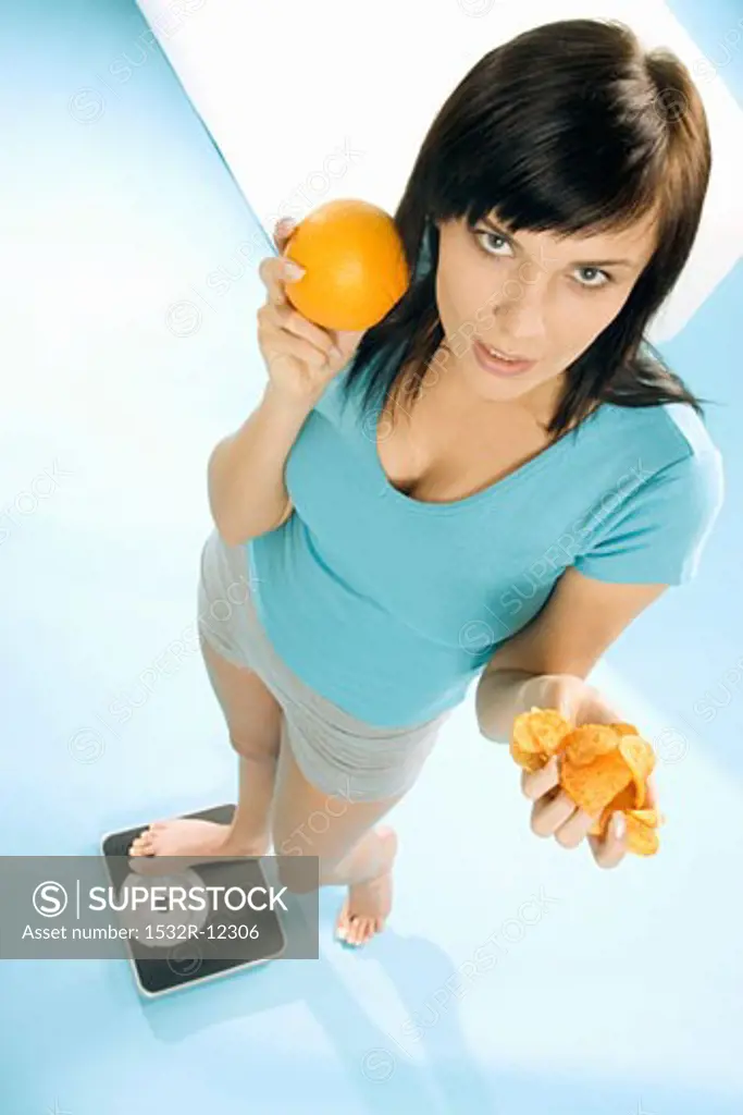 Young woman standing on scales with crisps and orange