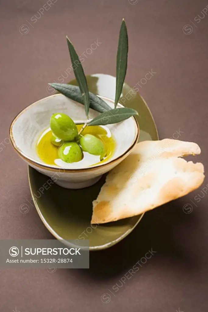Green olives on twig in bowl of olive oil, crackers