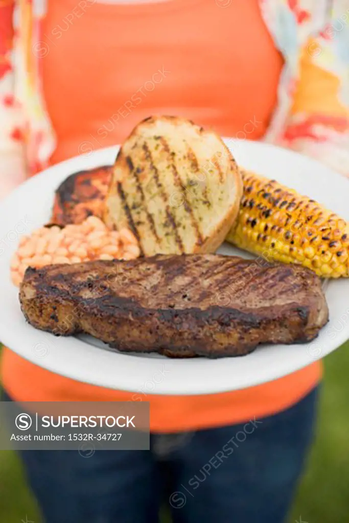 Woman holding plate of steak, bread, corn on the cob, baked beans