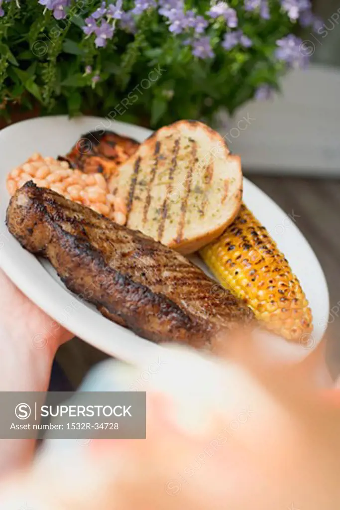 Hands holding plate of steak, bread, corn on the cob, baked beans