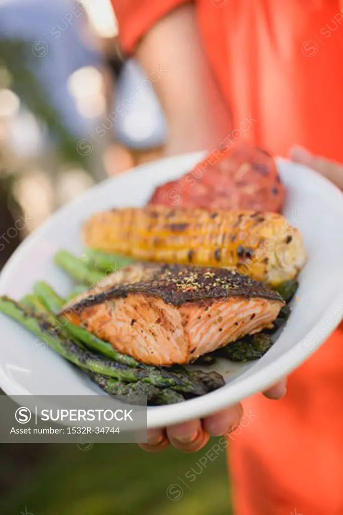 Woman holding plate of grilled salmon, corn on the cob & vegetables
