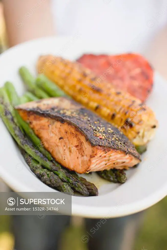 Person holding plate of grilled salmon, corn on the cob, vegetables