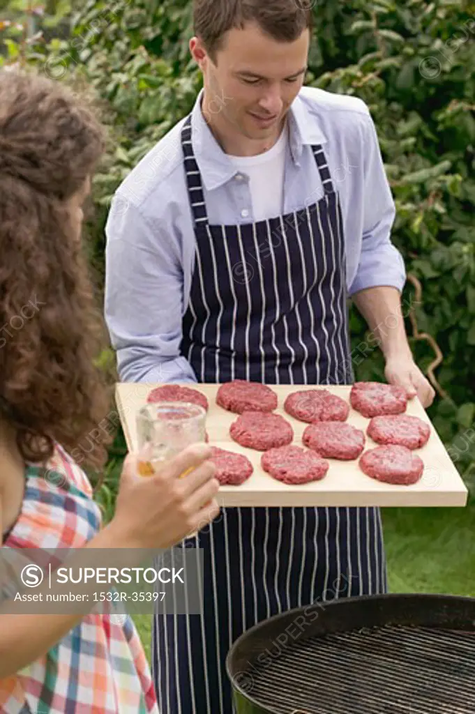 Man holding raw burgers over barbecue, woman holding iced tea
