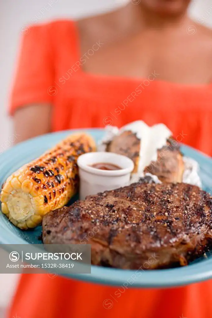 Woman holding plate of steak, corn on the cob, baked potato