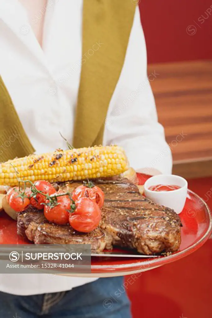 Woman holding T-bone steak, corn on the cob & cherry tomatoes