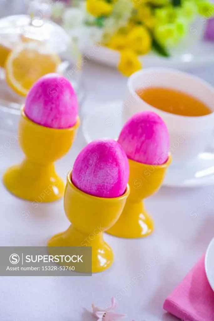 Pink Easter eggs in eggcups, cup of tea behind