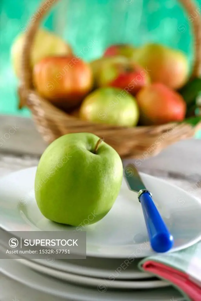 An apple on plate in front of a basket of fruit