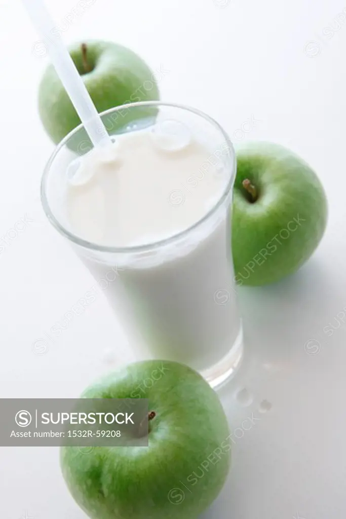 A glass of milk with a straw and three green apples