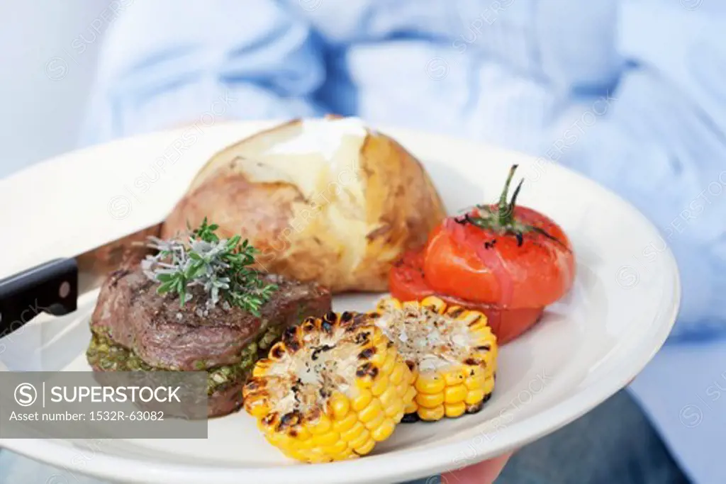 A person holding a plate of grilled beef medallions, a jacket potato and grilled tomatoes and corn on the cob