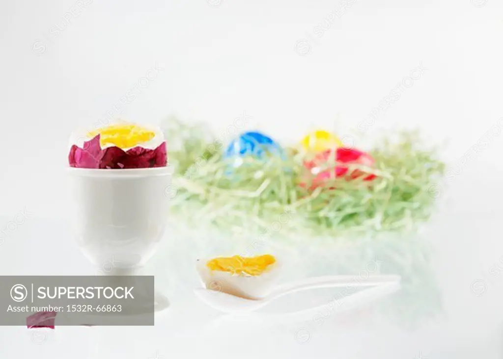 A boiled egg with the top cut off, in an eggcup, with an Easter nest of colourful eggs in the background