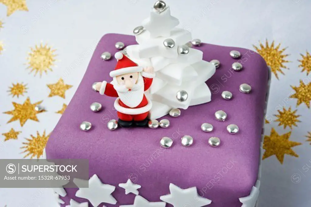 a model in icing of father Christmas holding up his arms on a purple and white Christmas cake with white stars and golden stars on a white background.