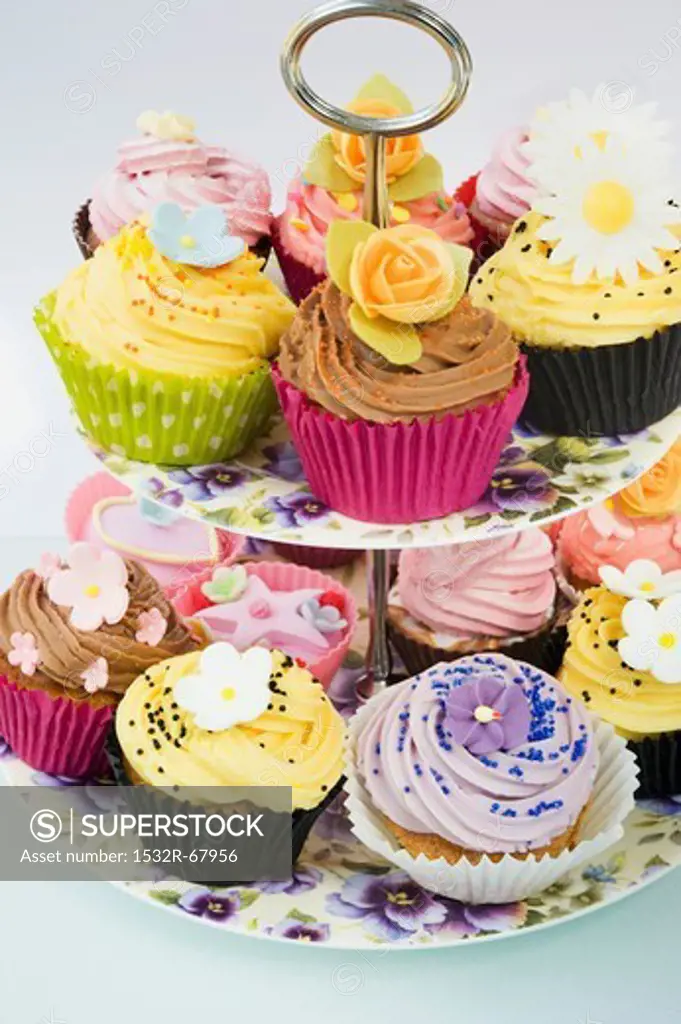 close up of a selction of iced purple, yellow and pink cup cakes decorated with icing flowers on a purple pansy flowered cake stand and a white background