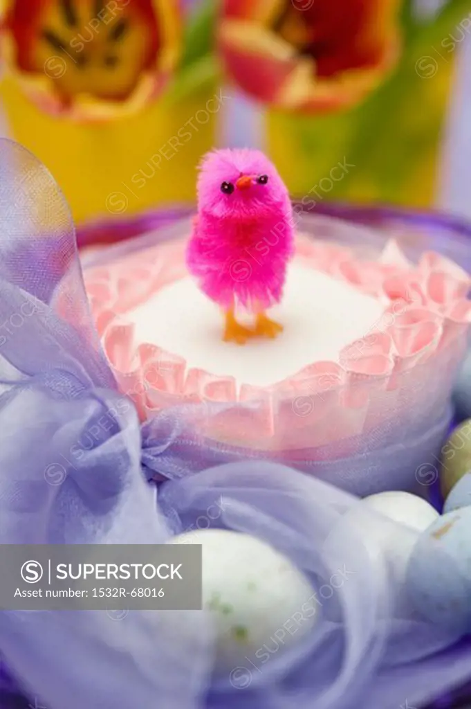 close up of single pink chick on top of a square easter cake tied with a purple chiffon bow surrounded by chocolate eggs and red and yellow tulip flowers