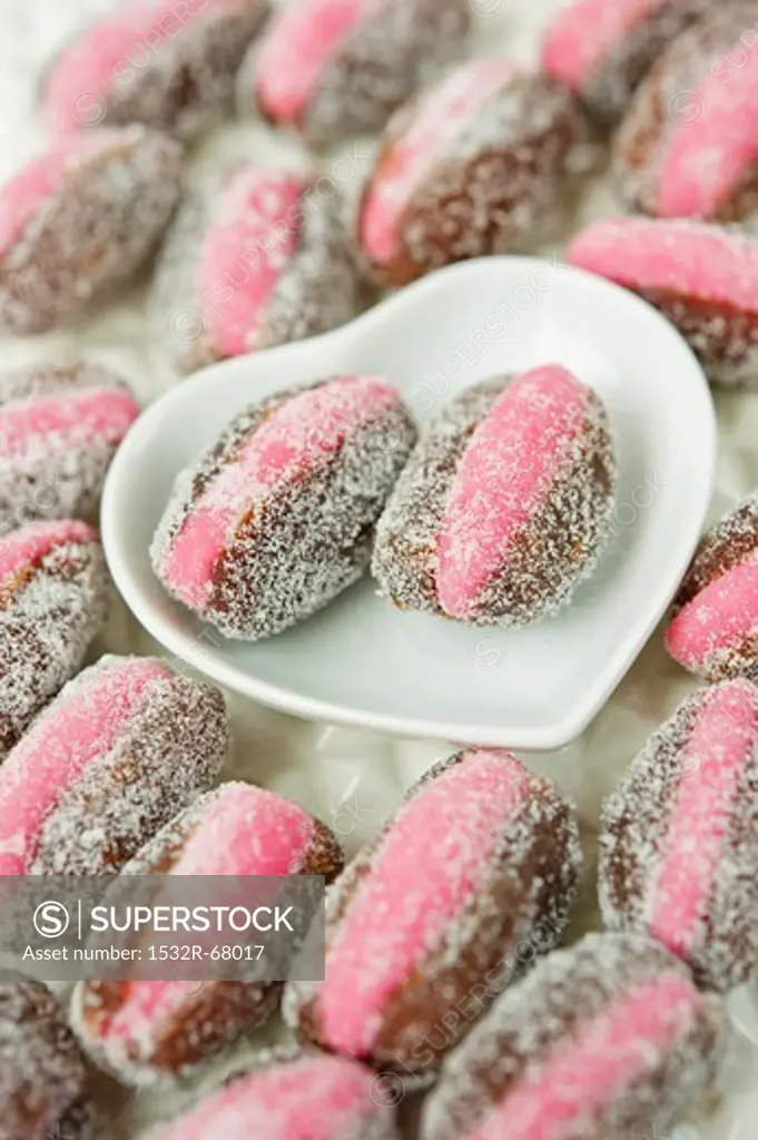 close up of pink marzipan stuffed dates with two laying in a white heart shaped ceramic dish