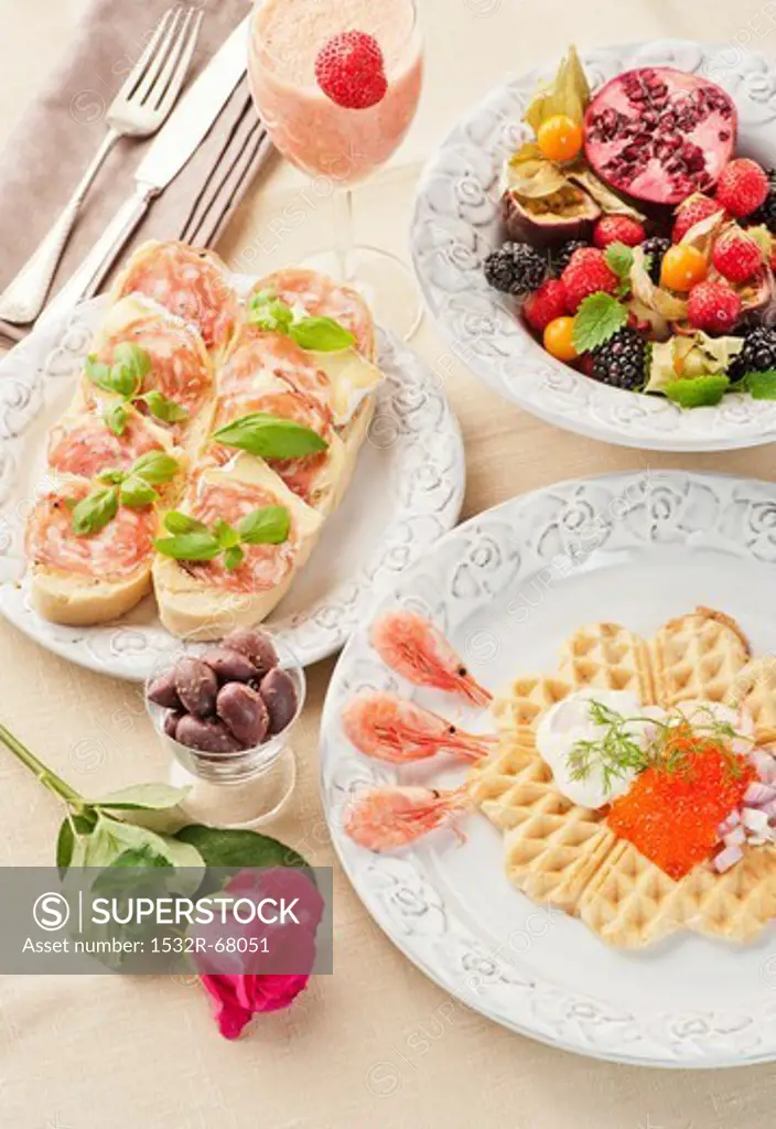 A breakfast of a caviar-topped waffle, slices of bread topped with cheese and ham, fresh fruits and a strawberry and mango smoothie