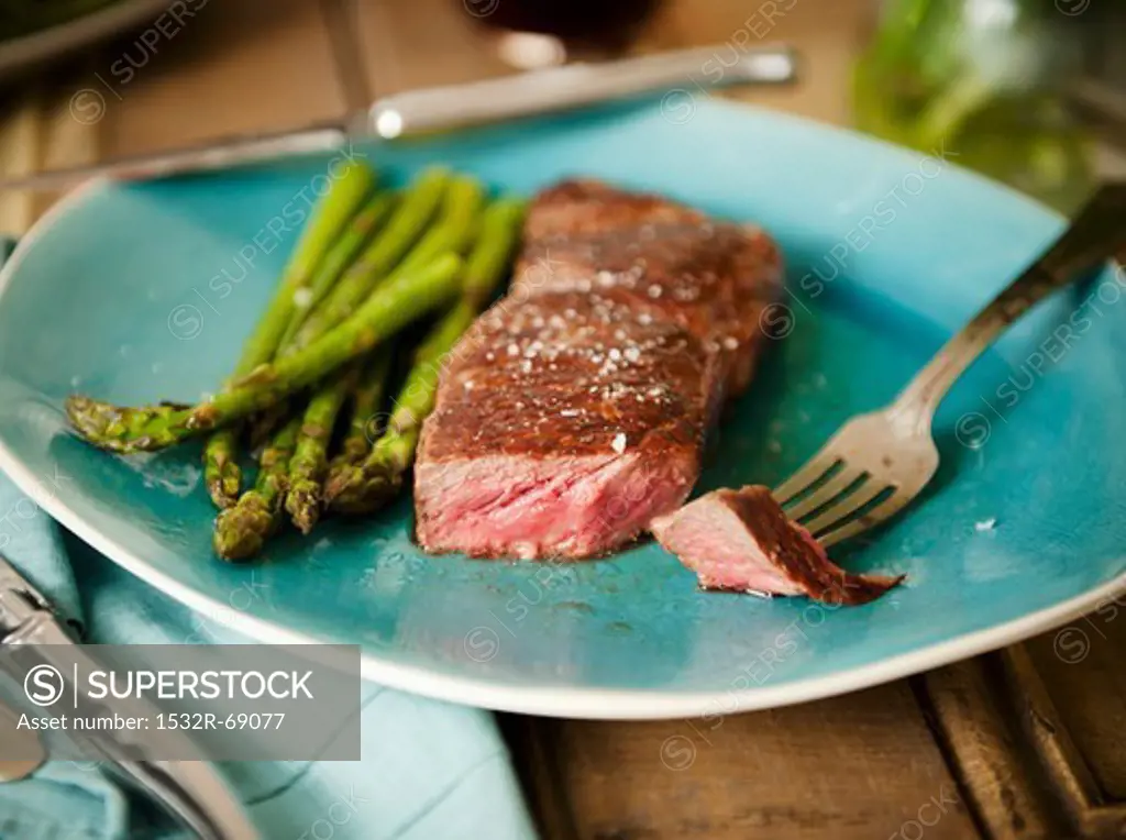 Strip Steak with a Piece Pierced on a Fork; Served with Asparagus on a Blue Plate