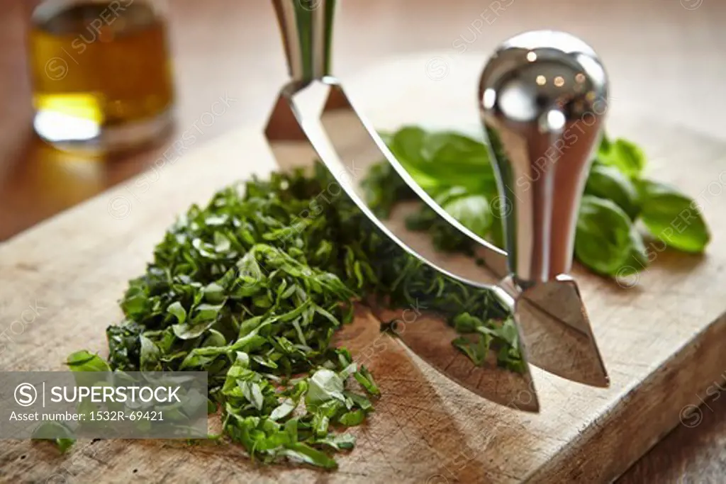 Basil being chopped with a curved chopping knife on a wooden board