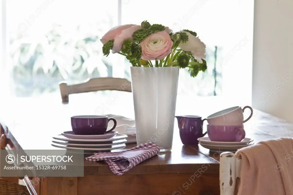 A wooden table with stacked crockery; in the centre is a decorative bunch of flowers including ranunculus