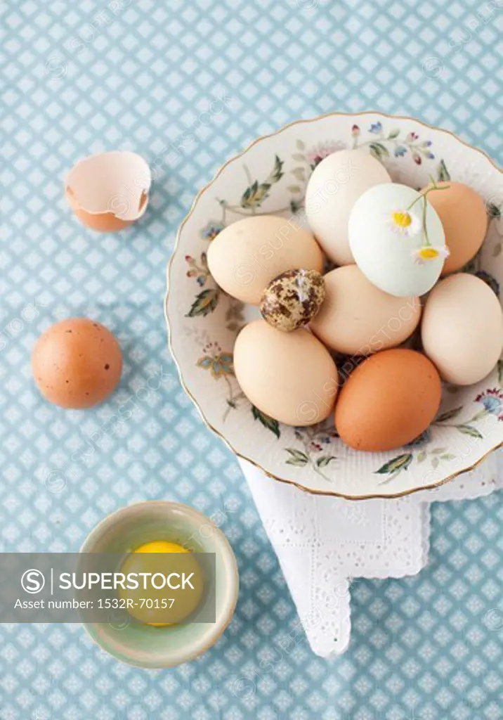 A Variety of Eggs in a Bowl with a White and Yellow Flower; From Above; One Egg Cracked Open