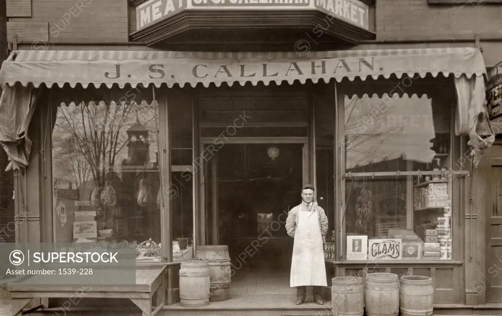 Shop owner stands outside of J.S. Callahan butcher shop general store postcard c. 1910