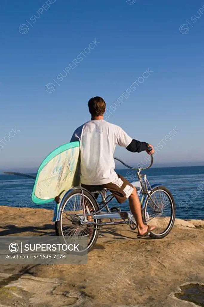 Rear view of a young man riding a bicycle and carrying a surfboard