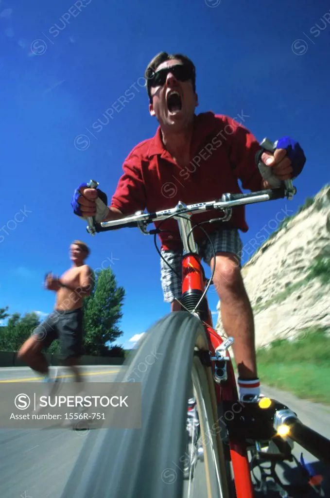 Low angle view of a young man riding a cycle with another young man running behind him