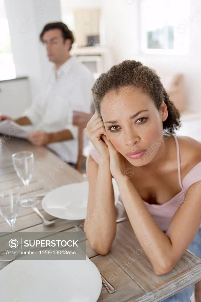 pensive young woman at the table, looking angry in the camera, man blurred in the background