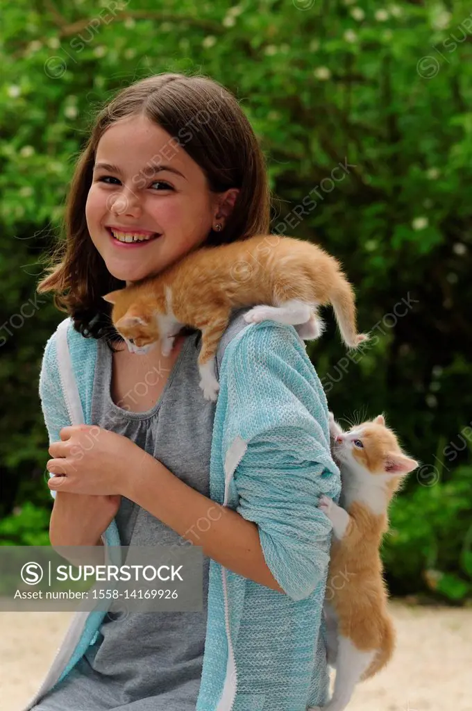 Girl with two kittens on shoulder and back laughs in the camera, trees in the background, blurred