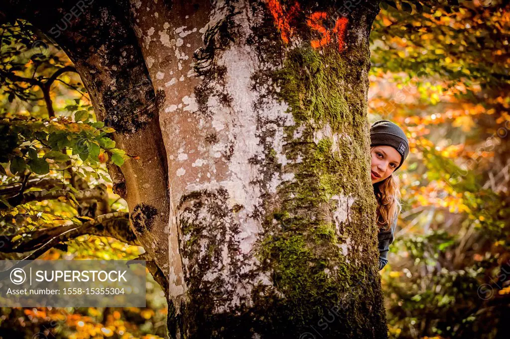 Young girl in autumn forest looking from behind tree into the camera