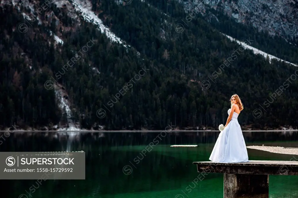Young blond woman in bridal gown on a footbridge at the lake with mountains in the background, daylight