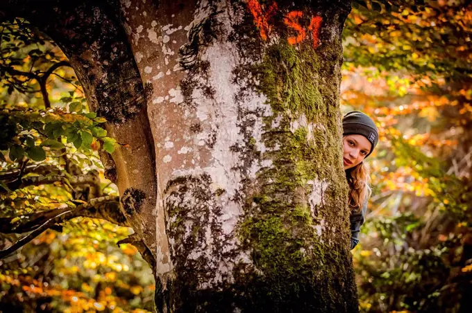 Young girl in autumn forest looking from behind tree into the camera