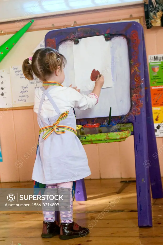 Rear view of a 3 year old girl, painting at nursrey school, with an apron on, concentrating. Standing at an easel