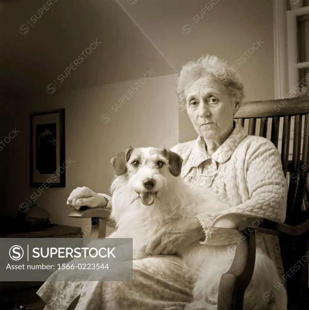 Concerne, worried looking elderly woman sitting on rocker in attic room (apparent from rooms ceiling line) with small dog on lap.  Both looking at cam...